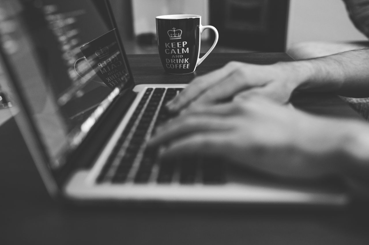 Monochrome image of hands on a laptop keyboard with a coffee mug, coding in progress.